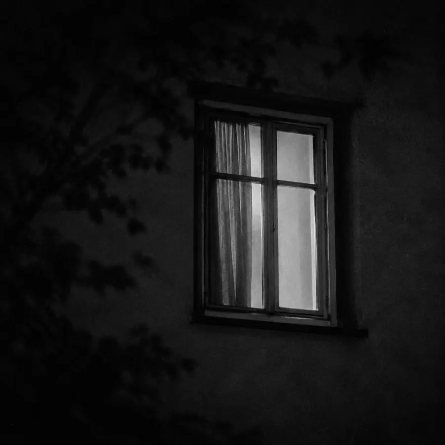 밤에 바깥에서 본 아파트 창문 하나. 커튼이 반쯤 쳐져 있고 실내 불빛이 밝은 회색으로 번진다.